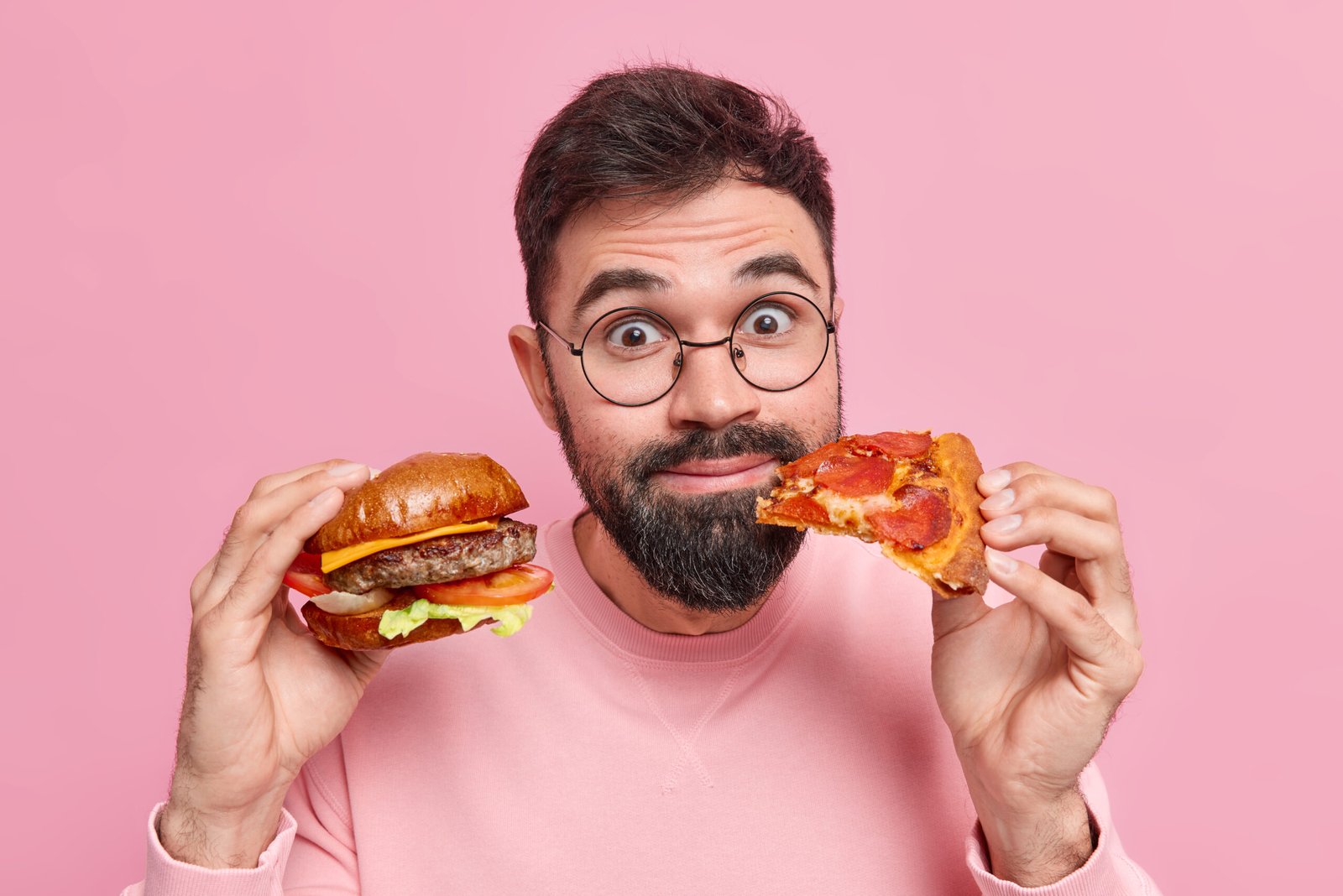 close up shot of surprised pleased bearded man holds burger and piece of pizza eats junk food doesnt care about health and nutrition wears spectacles neat jumper isolated over pink background